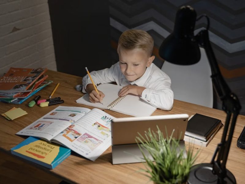 A young boy focused on studying at a wooden table with a tablet and books nearby under a desk lamp.