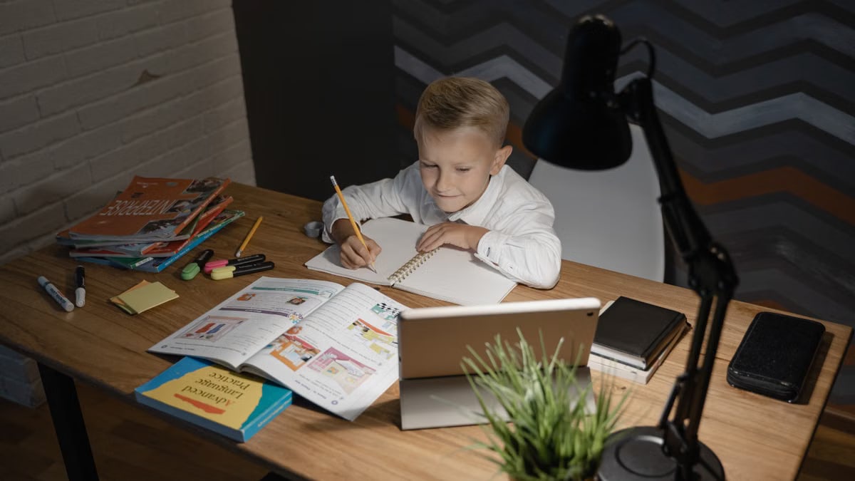 A young boy focused on studying at a wooden table with a tablet and books nearby under a desk lamp.